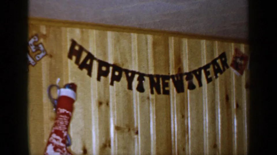 1965 Long Island home-movie frame with the HAPPY NEW YEAR banner centered over a red stocking on a paneled wall.
