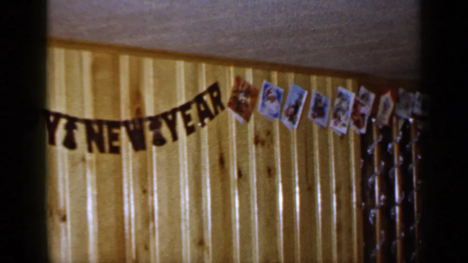 1965 Long Island interior with a Happy New Year banner, clipped holiday cards, and tinsel garland along a paneled wall.