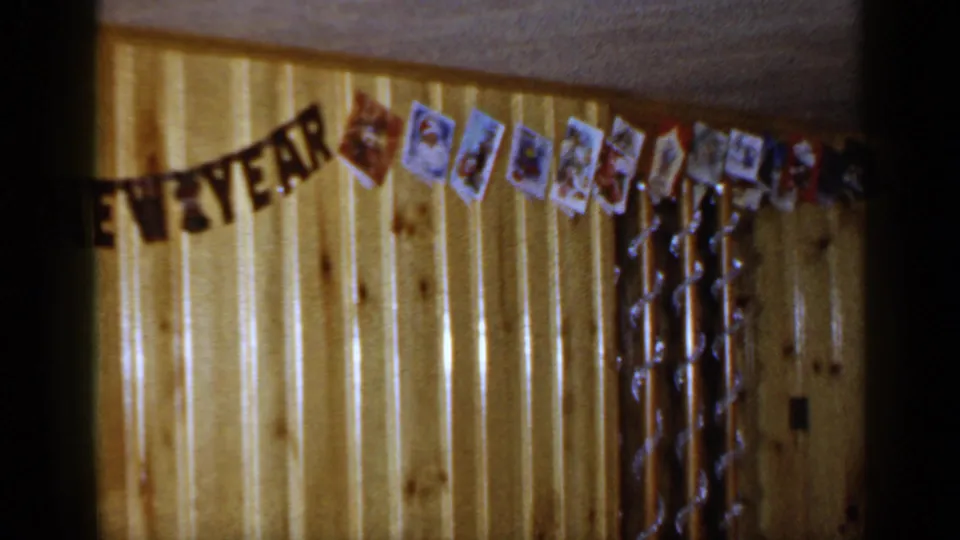 1965 Long Island home interior showing the end of a New Year banner with cards and tinsel on a wood-paneled wall.