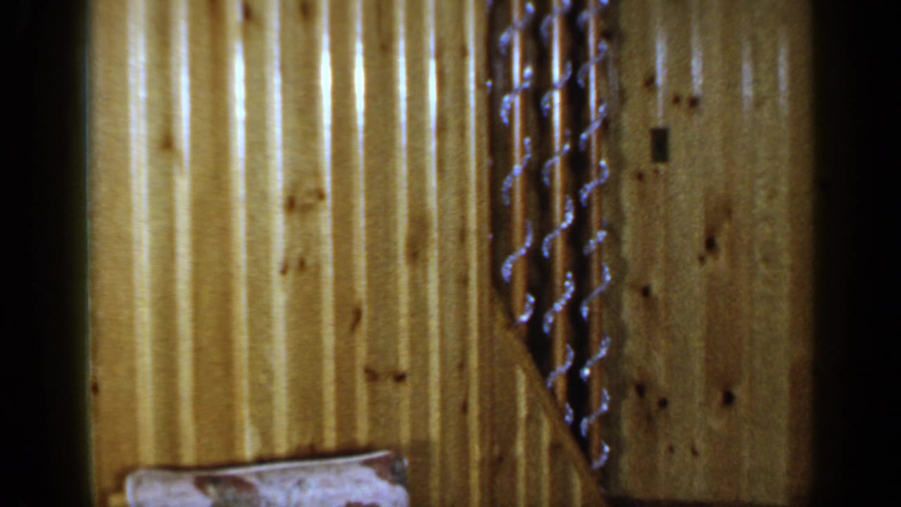 1965 Long Island home-movie frame showing knotty wood paneling, a stair rail, and a partial upholstered cushion in a private interior.