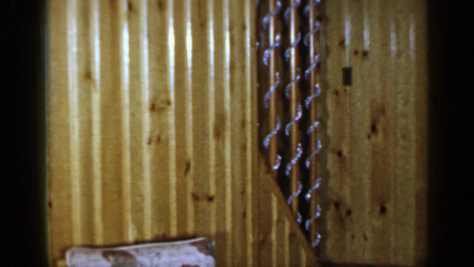 1965 Long Island home-movie frame showing knotty wood paneling, a stair rail, and a partial upholstered cushion in a private interior.