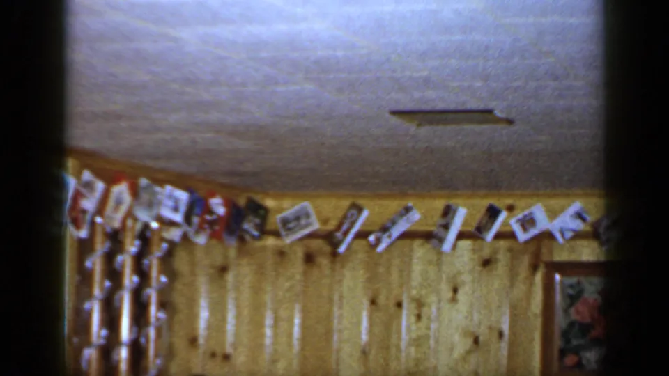 1965 Long Island interior view of cards or photos strung along a wood-paneled wall below a ceiling vent.
