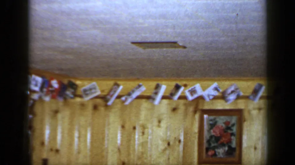 1965 Long Island home interior with a line of hanging cards or photos above a framed floral picture on wood paneling.