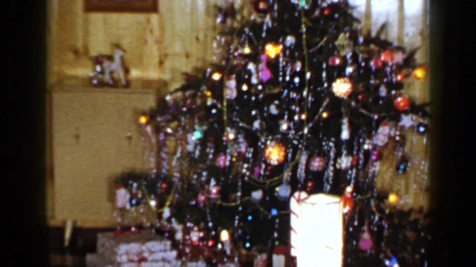 1965 Long Island home-movie frame focused on a tinsel-covered Christmas tree with colorful ornaments, lights, and a cabinet in the background.