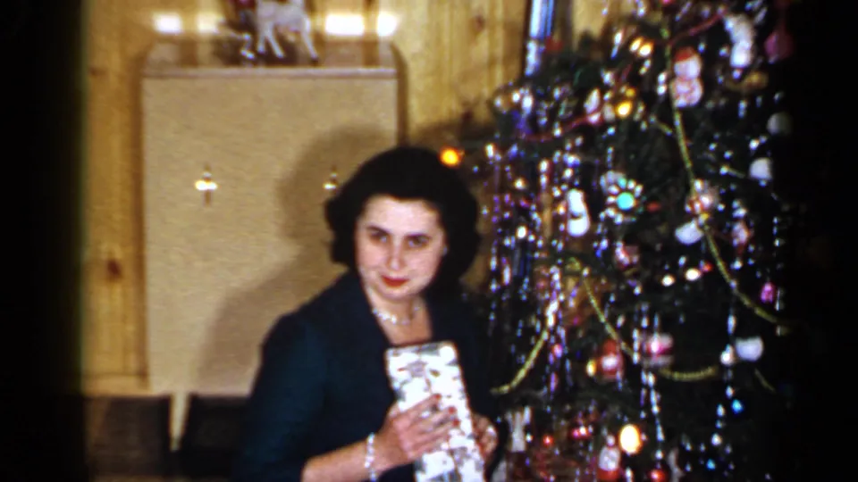 1965 Long Island home-movie frame showing a woman in dark clothing and pearls seated near a lit Christmas tree while holding a wrapped present.