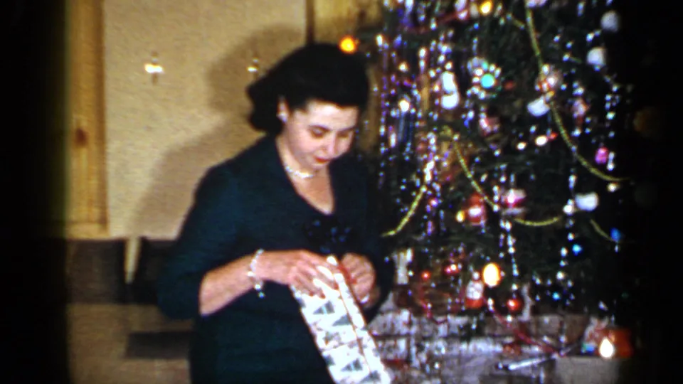 The woman sits near the lit and ornamented Christmas tree, looking down as she works at patterned wrapping paper.