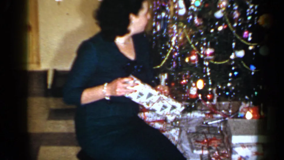 The woman turns toward the decorated tree with a gift in her hands, surrounded by ornaments, tinsel, and wrapped packages.