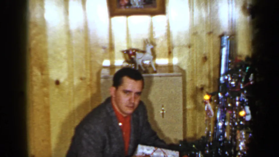 A man in a red shirt and jacket sits in the same wood-paneled room near the decorated Christmas tree, holding a wrapped gift.