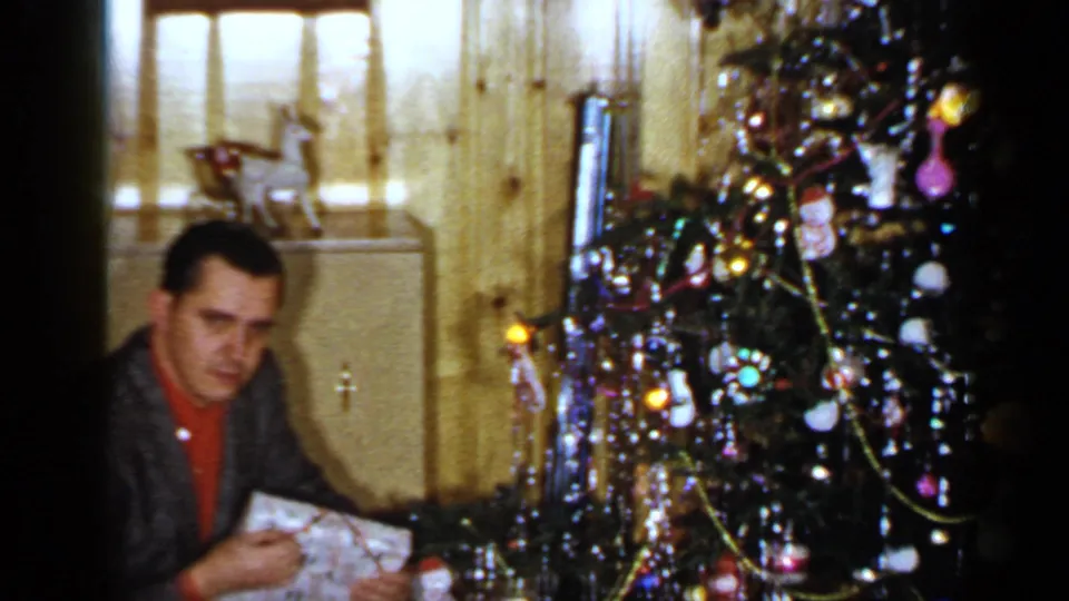 The man looks toward the camera beside the decorated Christmas tree in a wood-paneled Long Island room.