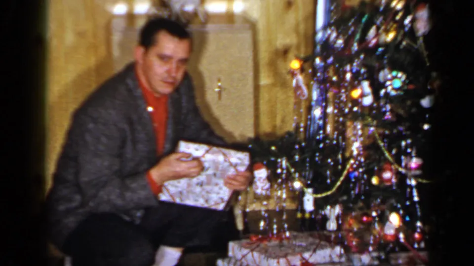 A kneeling man in a red sweater and gray jacket unwraps a gift beside the Christmas tree in the 1965 home-movie scene.