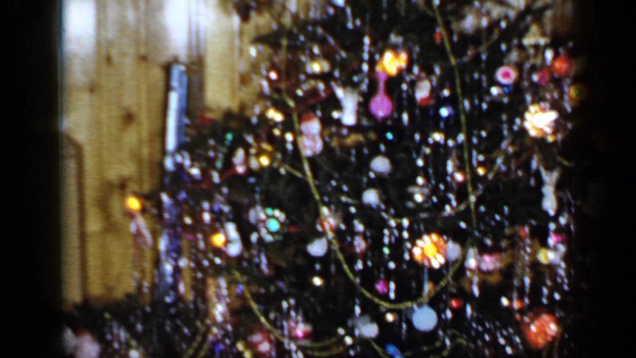 Close 1965 Long Island home-movie view of a Christmas tree covered with tinsel, ornaments and lights inside a wood-paneled room.