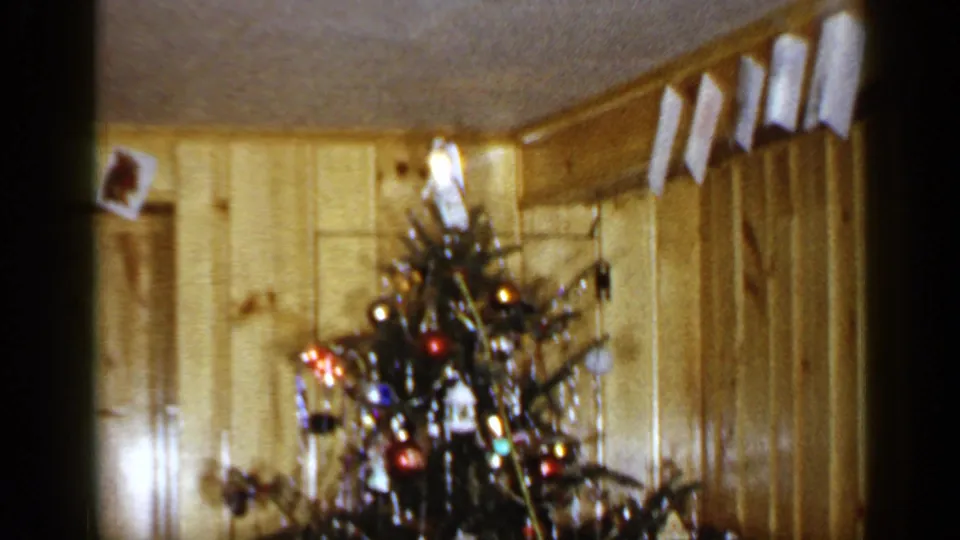 Wider 1965 Long Island home interior showing a decorated Christmas tree below hanging cards in a wood-paneled living room.