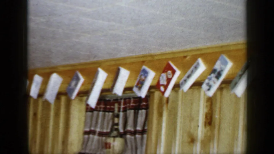 The camera frames a row of Christmas cards hanging near the ceiling over wood paneling and a small curtain in the 1965 home interior.