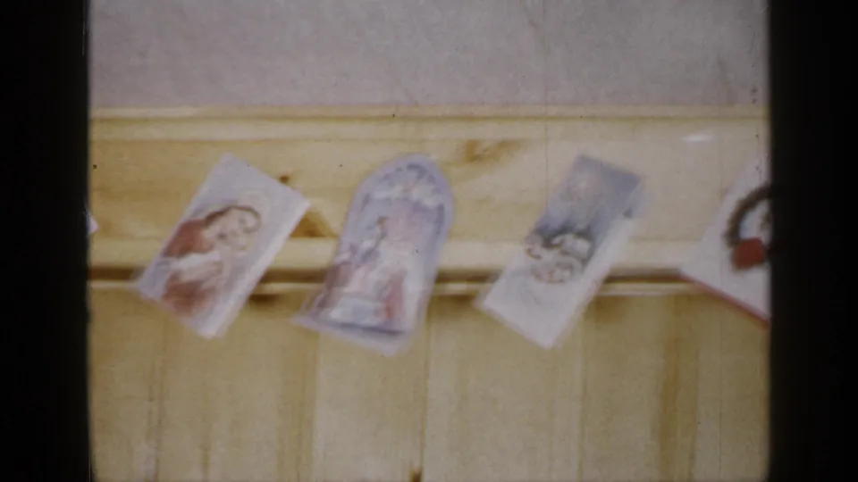 Religious greeting cards, including saint or Madonna imagery, hang from a line across a wooden wall in Long Island home-movie footage.
