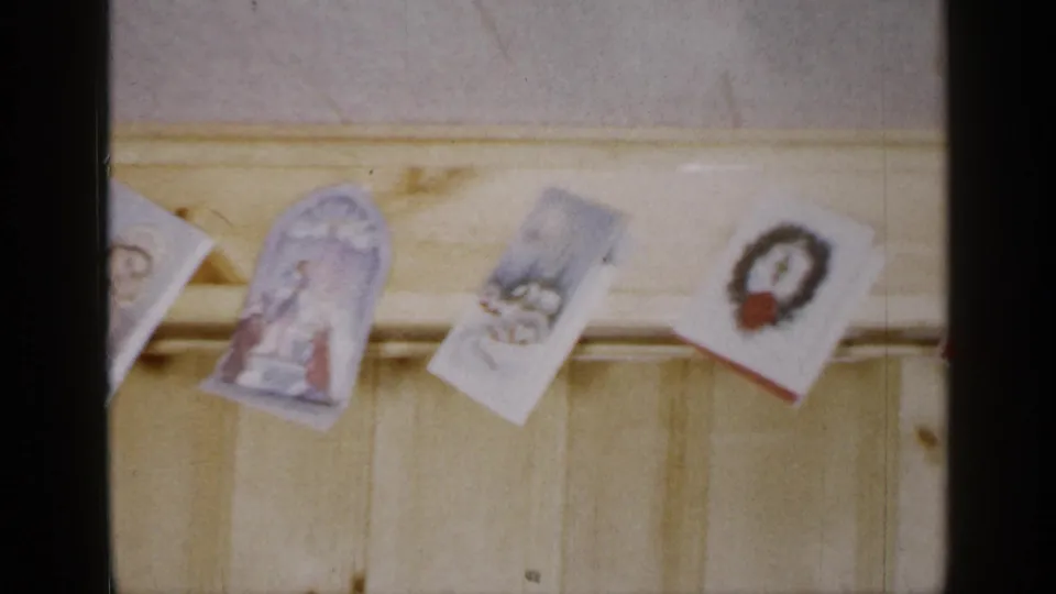 1965 Long Island home-movie frame showing Christmas cards clipped or strung along a pale yellow wooden beam inside a paneled room.