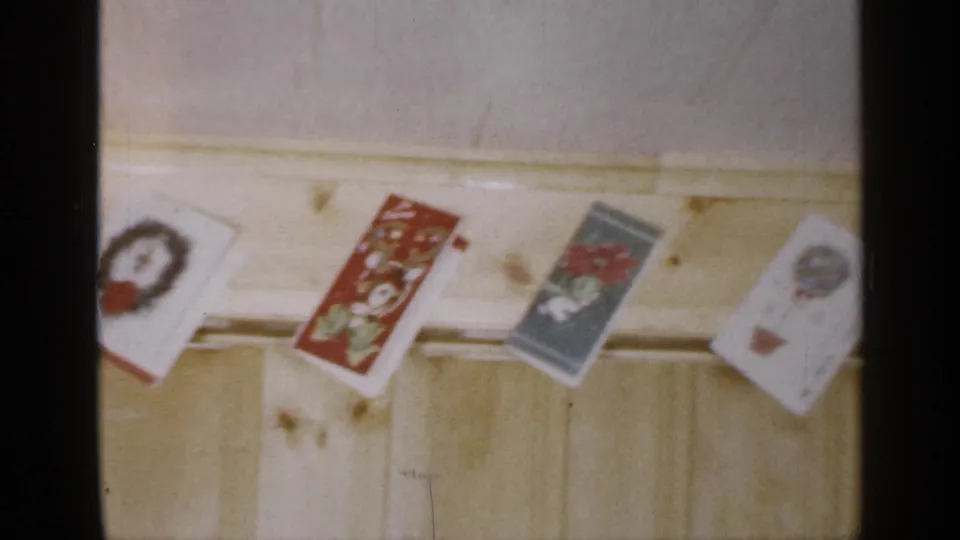 1965 8mm frame of Christmas cards hanging above pale paneling, including a wreath card, a red illustrated card, and a blue winter-themed card.