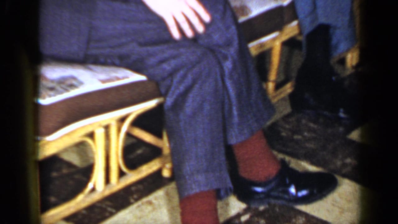 Close view of seated legs in dark trousers, red socks, polished shoes, and wicker chairs.