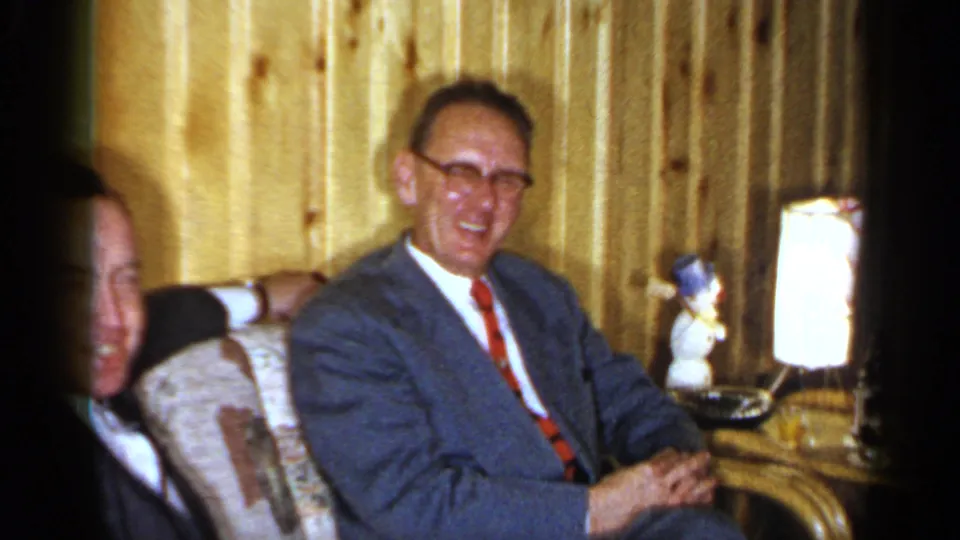 1965 Long Island home-movie frame of a smiling man in glasses, blue suit, and red tie seated beside another adult in a wood-paneled interior.
