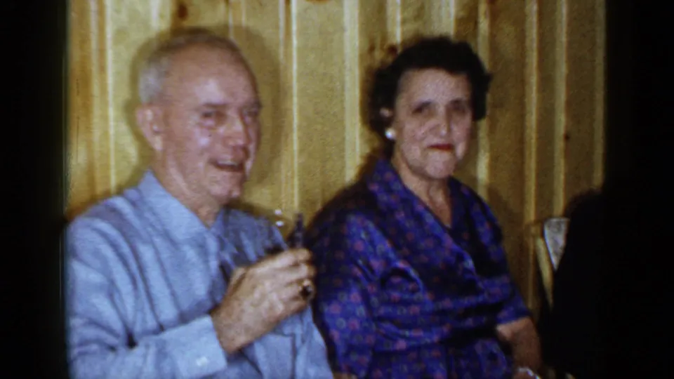 1965 Long Island interior frame showing an older man with a drink seated next to a woman in a patterned blue dress against wood paneling.