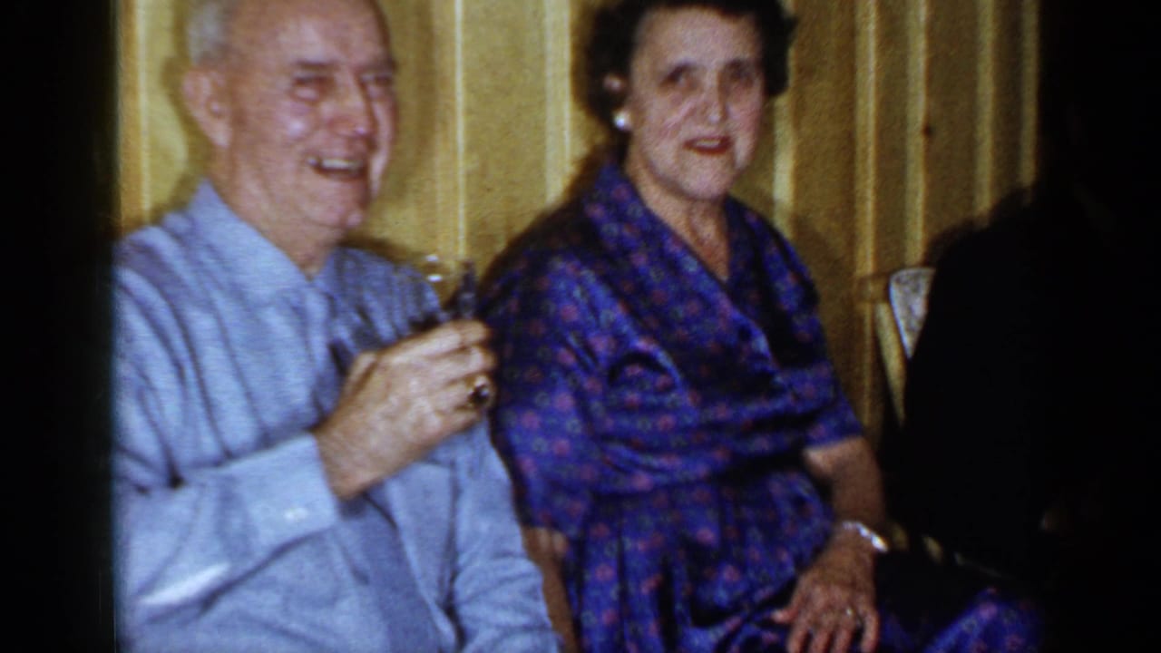 Elderly couple seated indoors, with the man smiling and holding a small glass.