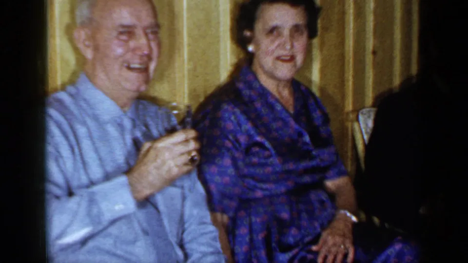 1965 Long Island home-movie frame of an elderly couple seated by a paneled wall during an indoor social gathering.