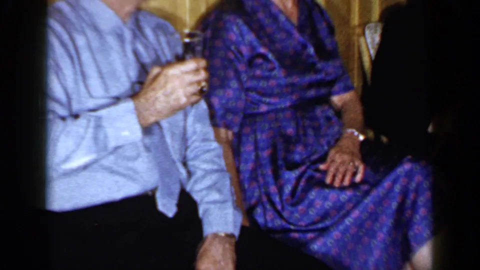Cropped 1965 Long Island home-movie frame showing the seated couple from chest to lap in an indoor setting.