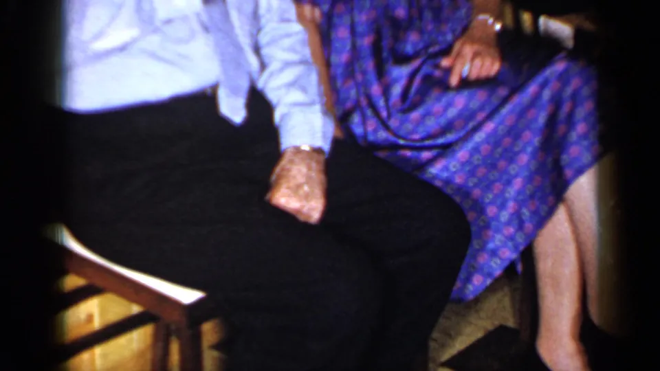 1965 Long Island home-movie frame focused on the seated couple's laps, chair arms, and checkered floor indoors.