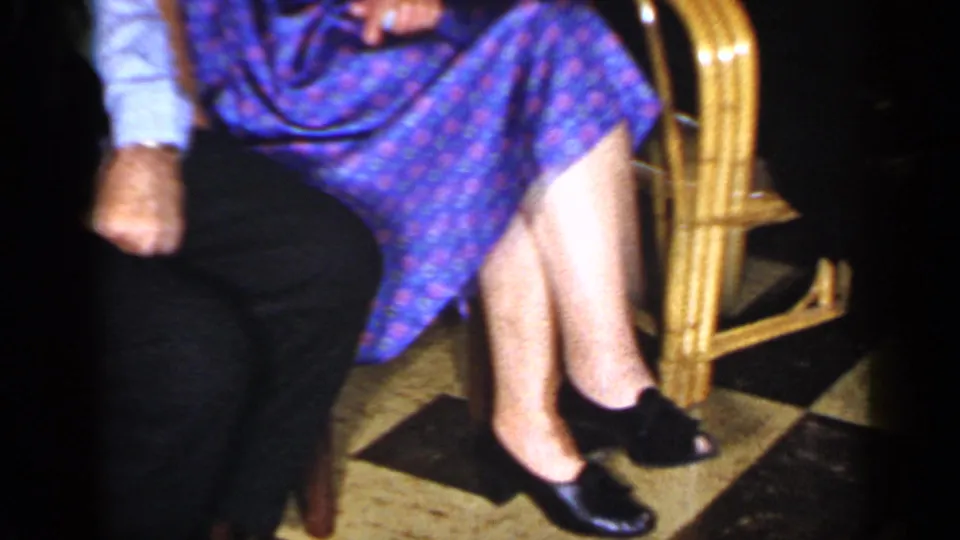 Cropped 1965 Long Island home-movie frame of the woman's dress, legs, shoes, and chair beside the seated man.