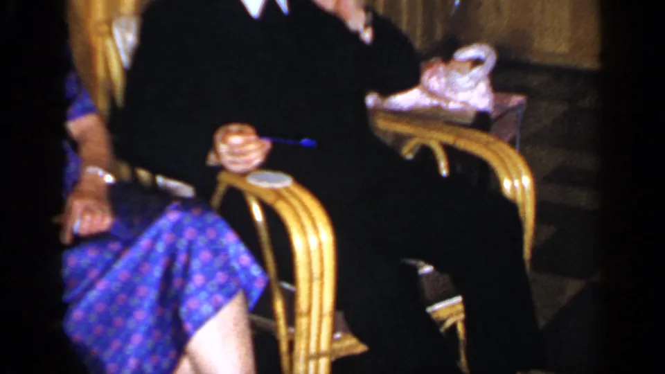 1965 Long Island interior with seated adults in wicker chairs, a woman in a patterned dress, drinks, and gold curtains in view.