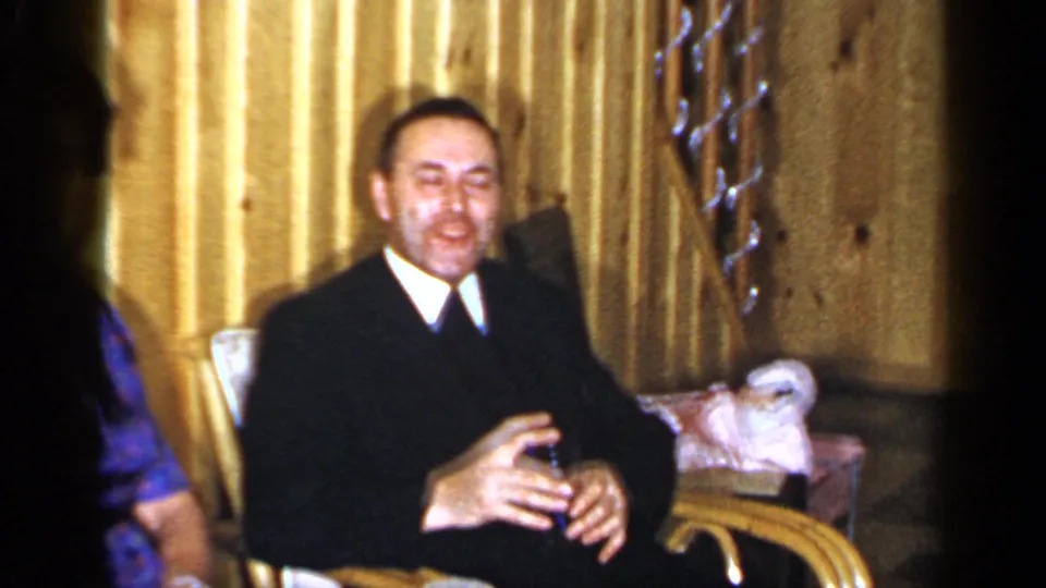 1965 Long Island frame of a smiling man in a dark suit seated in a wicker chair, holding a drink against gold curtains.