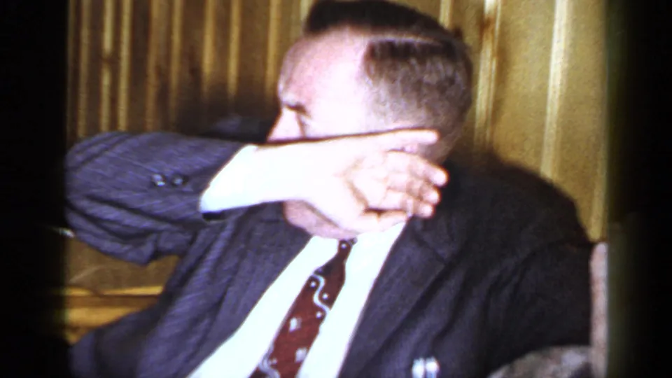 1965 Long Island home-movie frame showing a suited man seated indoors, raising one arm across his face near gold curtains.