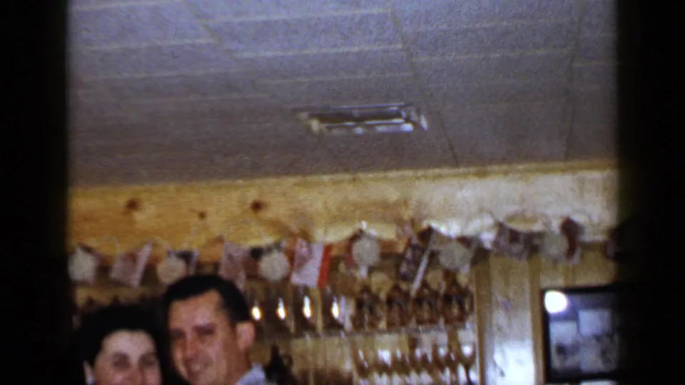 1965 Long Island home-movie frame with a smiling couple partly visible below ceiling tiles, garland, and bar glassware.