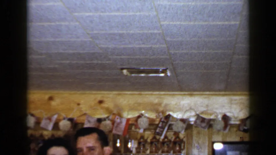 1965 Long Island bar interior with the couple low in frame, hanging decorations, glasses, bottles, and a framed item on the wall.