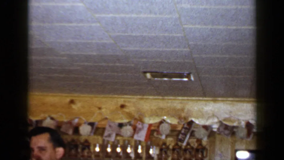 1965 Long Island home-movie frame showing a man cropped at the lower edge beneath ceiling tiles and a decorated bar wall.