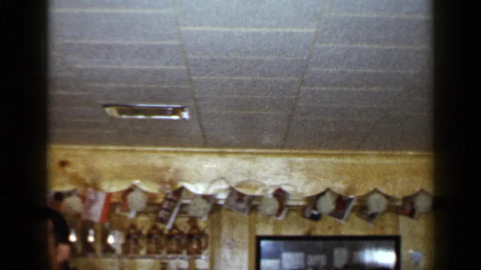 1965 Long Island bar scene with a partial adult face, garland, glassware, bottles, ceiling tiles, and framed wall decor.