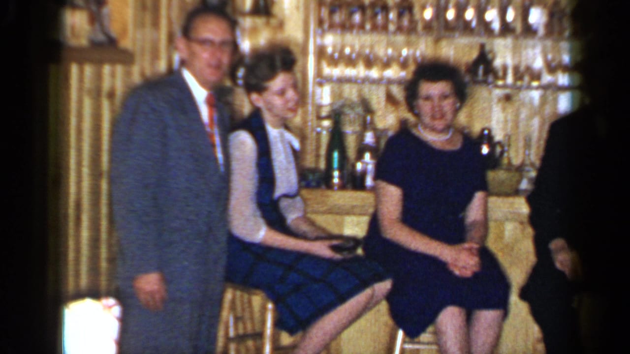Man in a suit stands beside two women seated at a wood-paneled bar.