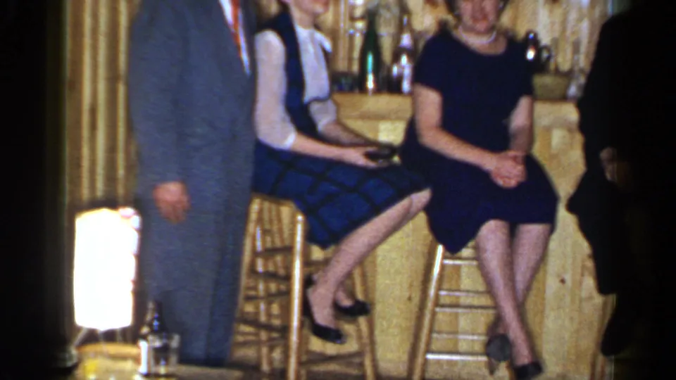 1965 Long Island frame showing two seated women at a private bar, with a cropped suited man and shelves of glassware in the background.