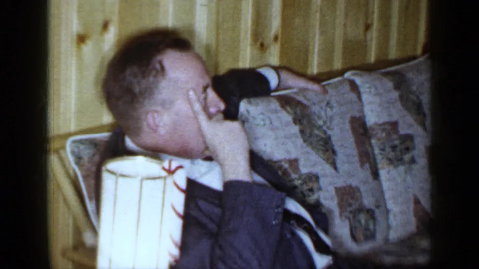 1965 Long Island home-movie frame of a man resting on a patterned sofa in a warm wood-paneled private interior.