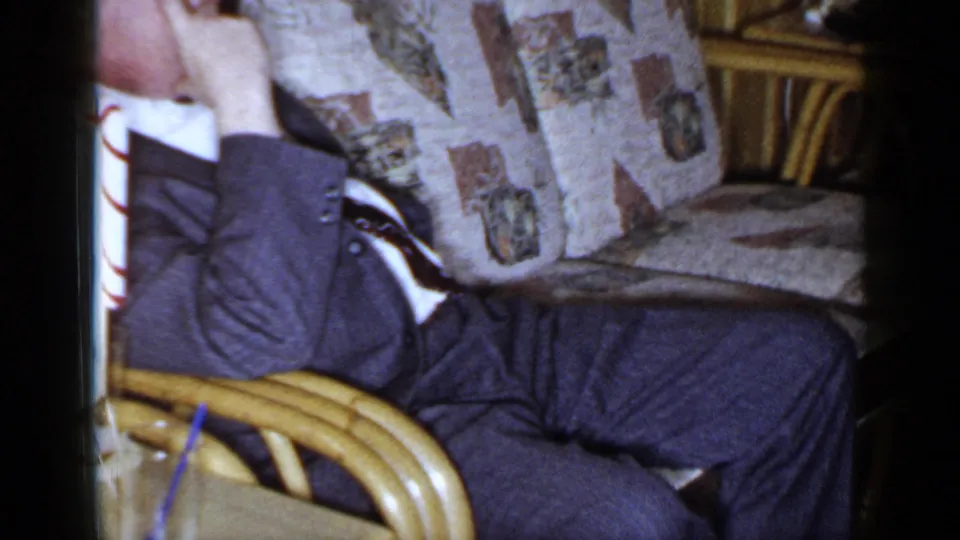 Cropped 1965 Long Island frame of a man reclining on a patterned sofa, partly obscured by a chair arm in a private interior.