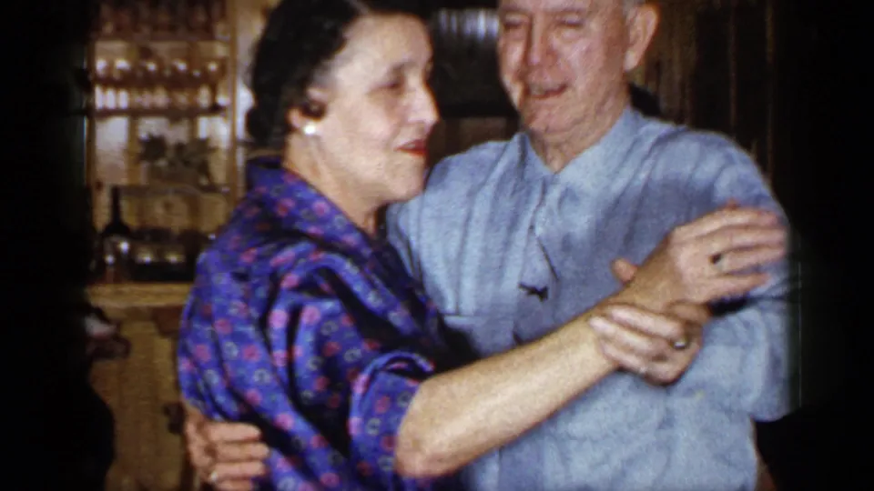 The older couple continues dancing in the Long Island interior, with the woman in a purple patterned dress and the man in a pale blue shirt.