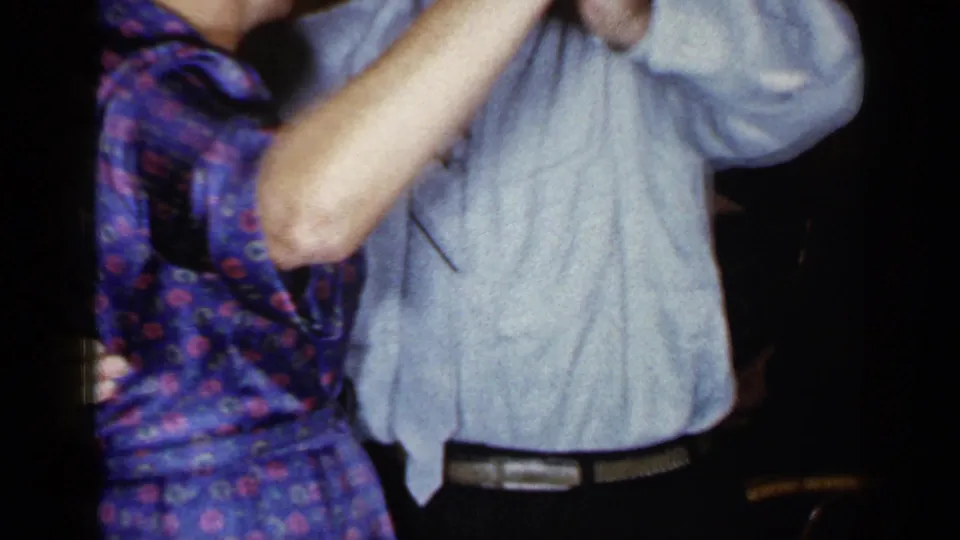 A close 8mm frame shows the couple mid-dance, their clasped hands and clothing dominating the warm indoor scene.