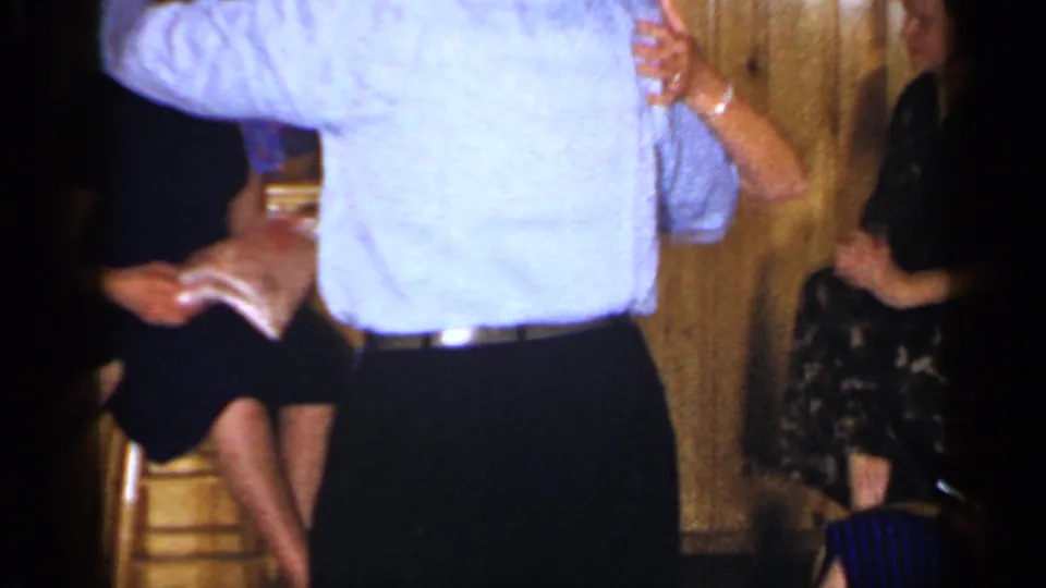 1965 Long Island home-movie frame of a couple dancing close to seated guests in an indoor gathering space.