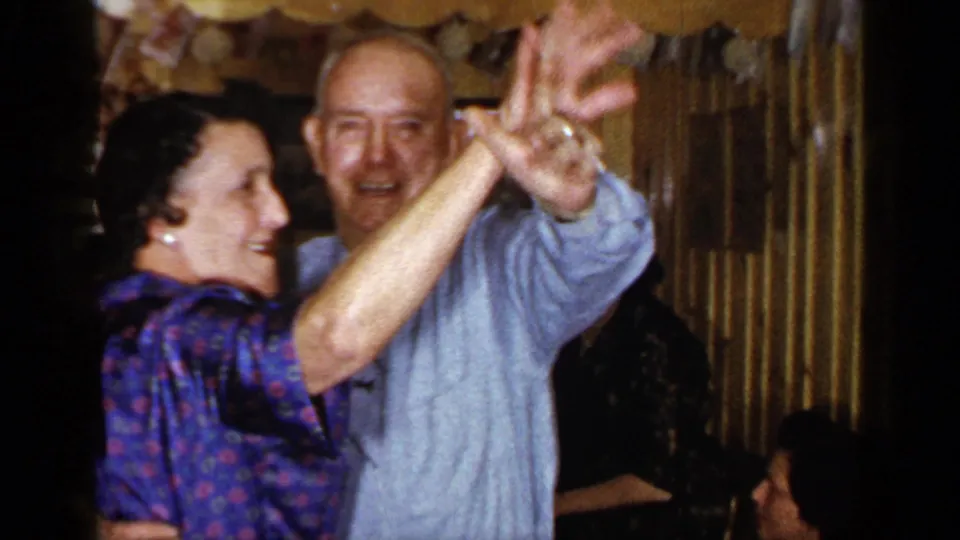 1965 Long Island home-movie frame of a smiling elderly couple dancing and appearing to wave toward the camera.