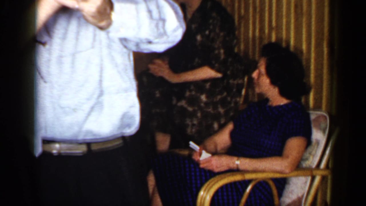 A man in a light shirt dances in front of women seated and standing in a Long Island bar interior, filmed in 1965.