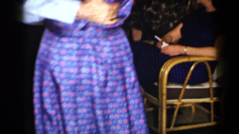1965 home-movie frame with a patterned dress in the foreground and a seated woman holding a cigarette beside rattan bar seating.