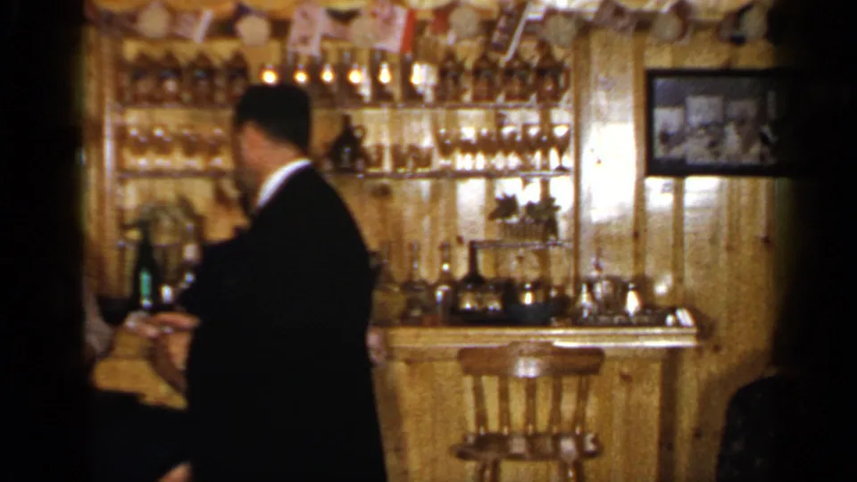 A man in a dark jacket appears beside a wooden bar with shelves of bottles and glassware in a 1965 Long Island interior.