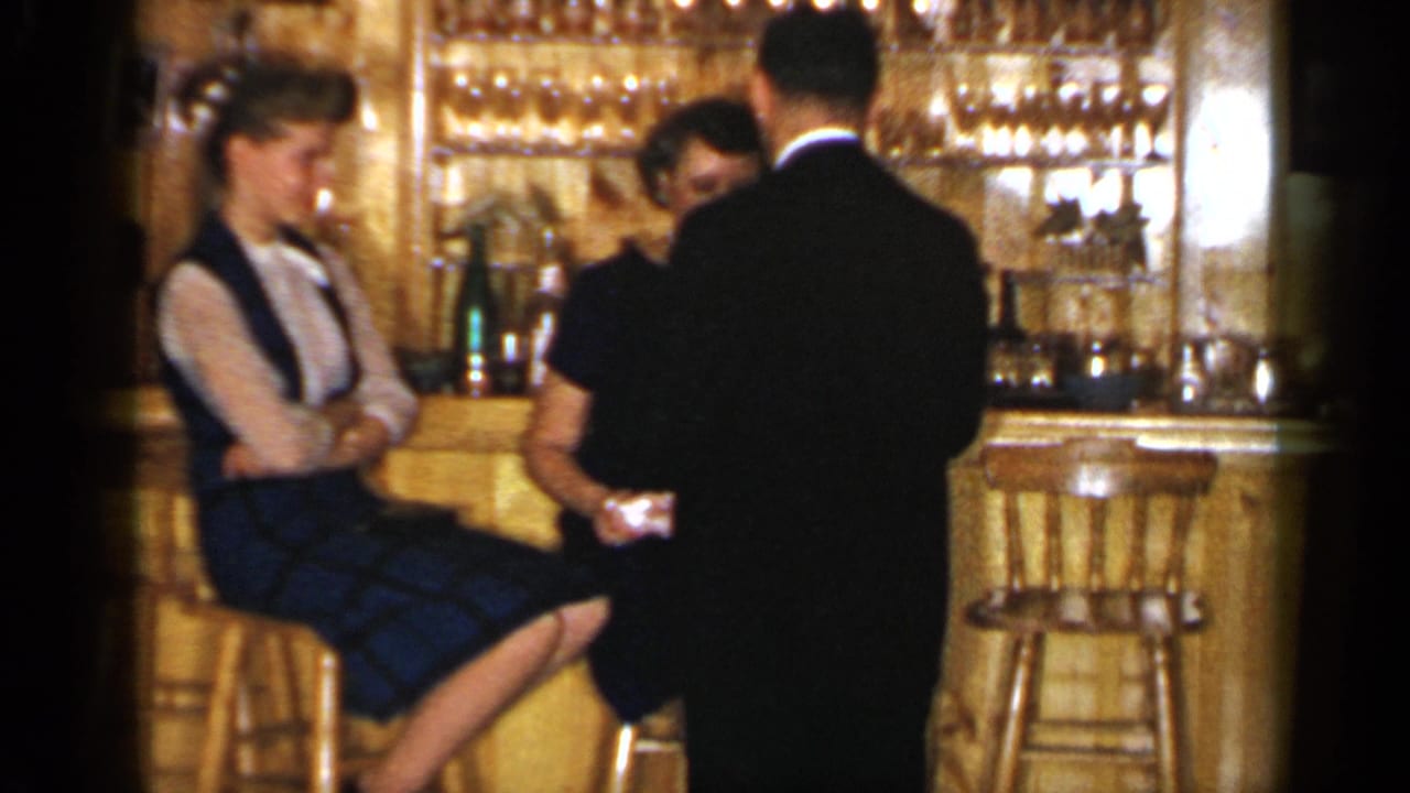 Two women and a suited man at a wooden bar with glassware in the background.