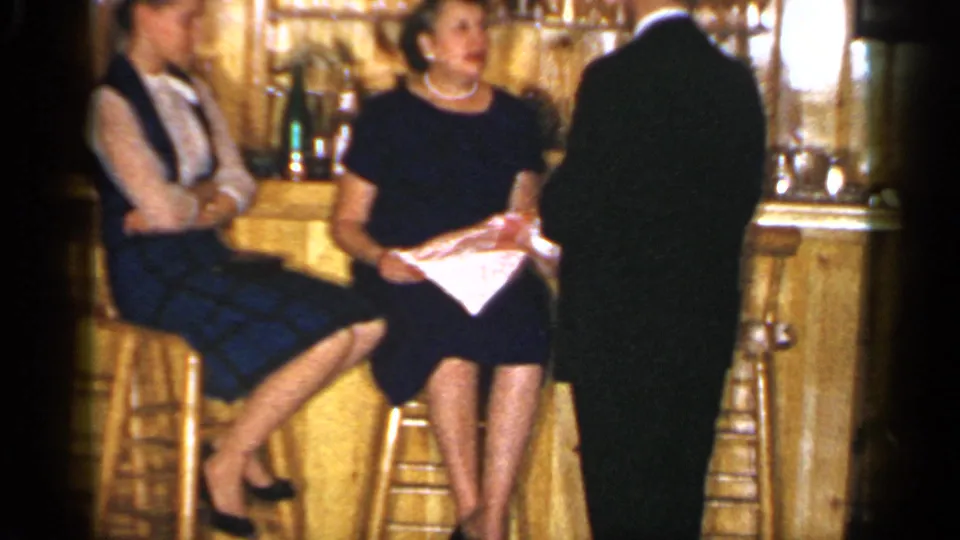 1965 Long Island home-movie frame showing two women seated on stools as a suited man stands with his back to the camera.