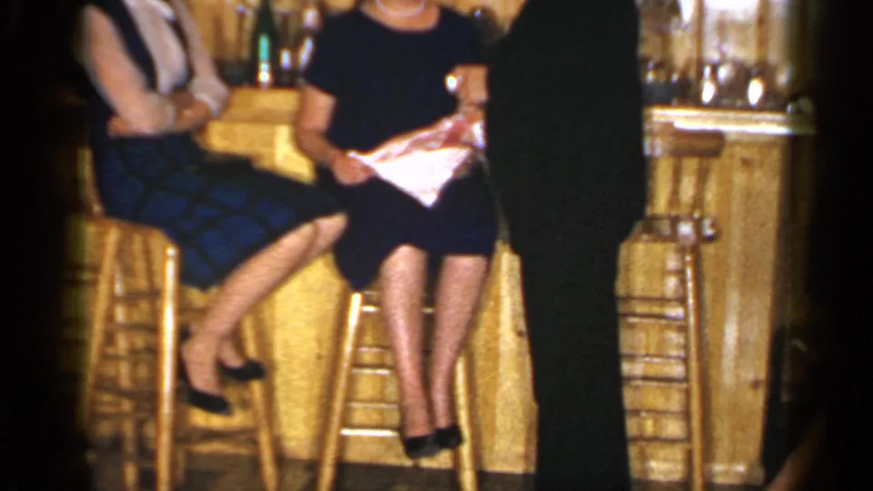 1965 Long Island home-movie frame of a suited man and seated women in conversation beside a rustic bar interior.
