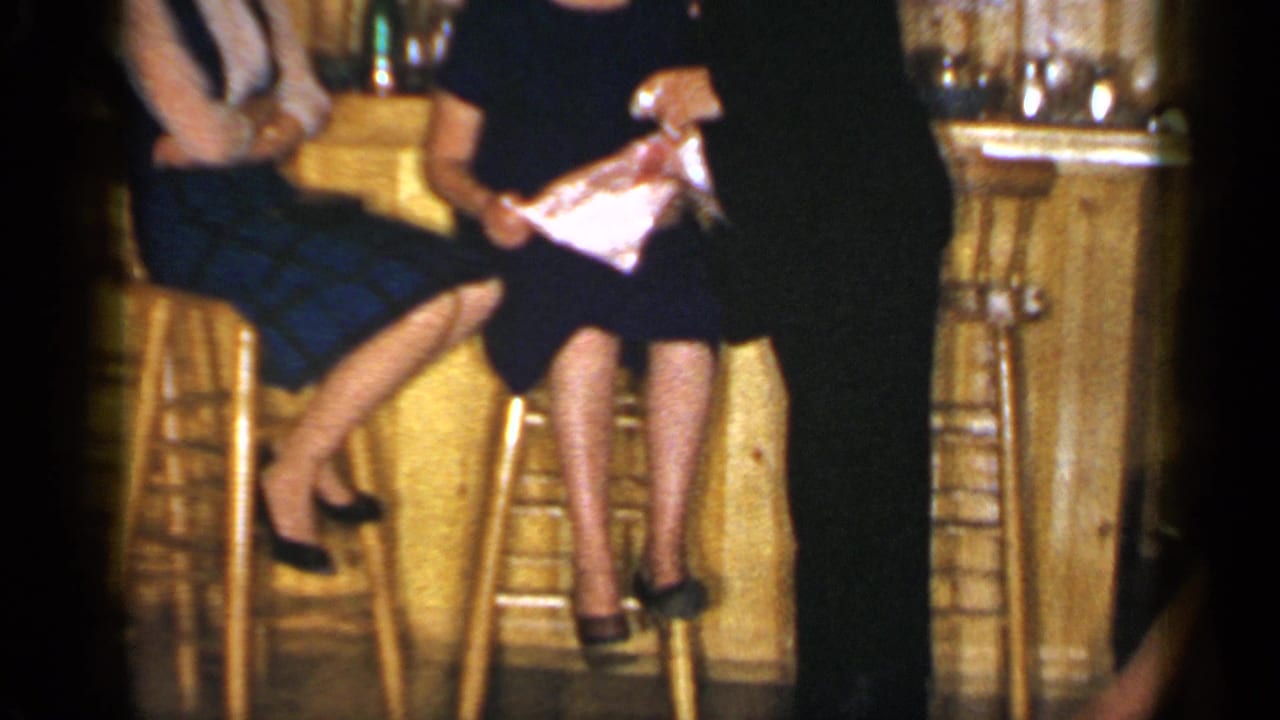 Two women sit on wooden barstools at a warm bar while a dark-suited man stands nearby.